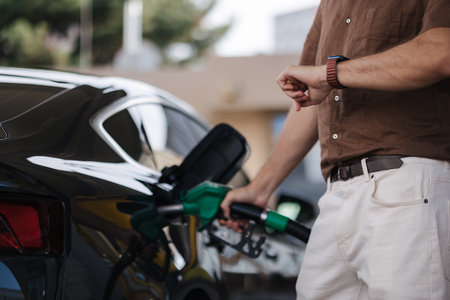 Low angle of male driver in casual clothes filling fuel into modern car with gas pump nozzle at station. Middle selection. Man looking at his watchの写真素材