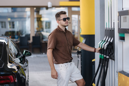 Man refueling a car at a petrol station. Guy in sunglasses hold fuel pump and look at the price of...