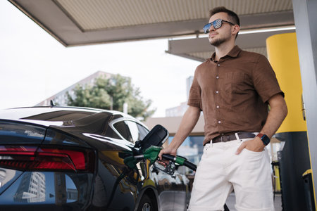 Low angle of concentrated young bearded male driver in sunglasses and casual clothes filling fuel into modern car with gas pump nozzle at station. Luxury carの写真素材