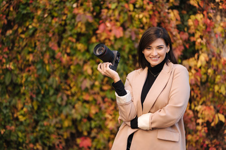 Happy smiled female photographer during her work. Stylish woman hold mirrorless camera. Autumn moodの写真素材
