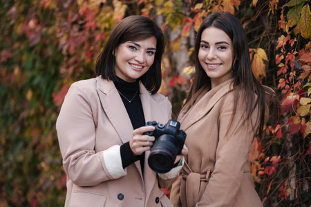 Girls are reviewing the photos they took on the camera. Two beautiful women stand in front of red and yellow trees.の写真素材