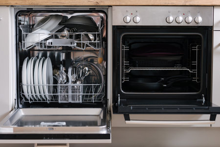 Open dishwasher placed beside an oven. Dishwasher loaded with clean dishes. Empty ovenの写真素材