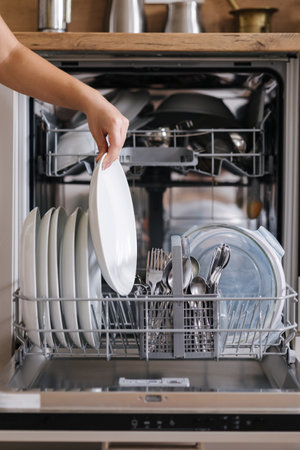 Front view from female hand removing clean plate from a loaded dishwasher. Background of modern kitchenの写真素材