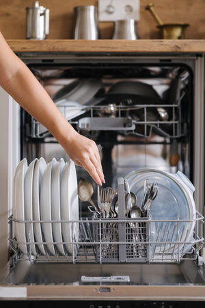 Front view from female hand removing clean cutlery from a loaded dishwasher. Background of modern kitchenの写真素材