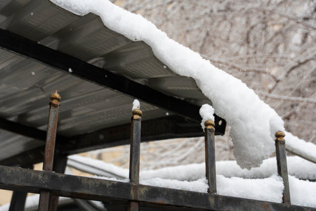 Metal fence and corrugated roof blanketed in thick snow, with bare trees in the backgroundの写真素材