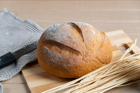 Golden-brown artisan bread with flour-dusted crust rests on wooden surface, accompanied by knife,...