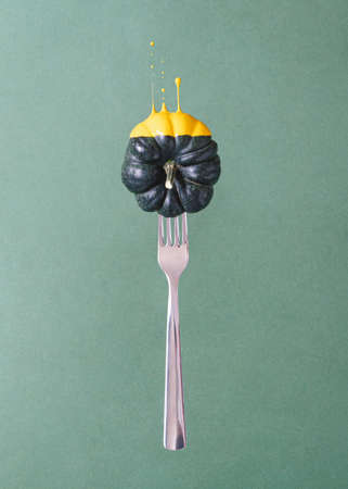 Creative composition with silver fork and dark pumpkin dipped in yellow liquid paint against green background.の写真素材