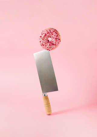 Surreal composition with tasty pink donut balancing on shiny meat cleaver
against pastel pink background.の写真素材