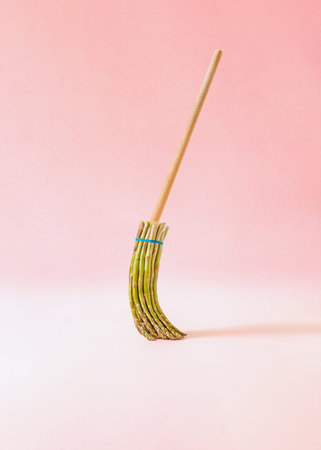 Creative surreal composition with broom made of fresh asparagus against pastel pink background....