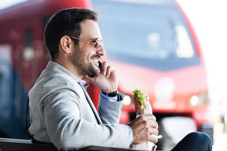 Young businessman on railroad station using phone and eating sandwich.の写真素材