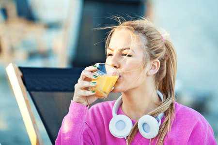 Blonde young woman drink orange juice after morning workout in cafe on the beach.の写真素材