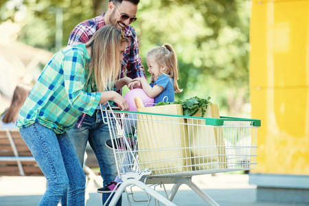 Young smiling parents and their cute daughter with shopping cart full of fresh food having fun in front of hypermarket.の写真素材
