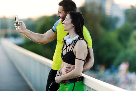 Early morning workout. Happy couple relaxing with cellphone after exercising on the bridge. Living healthy lifestyleの写真素材