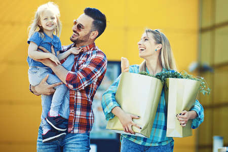 Young smiling parents and their cute daughter with shopping cart full of fresh food having fun in front of hypermarket.の写真素材