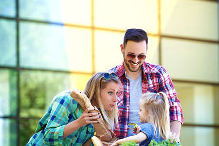 Young smiling parents and their cute daughter with shopping cart full of fresh food having fun in front of hypermarket.の写真素材