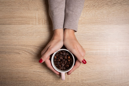 White Cup full of coffee beans in beautiful women's hands. Wooden background.の写真素材