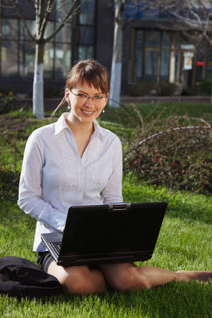 Woman lying on grass with laptop with business building on backの写真素材