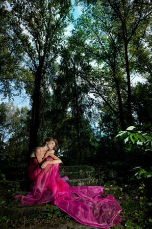 Gentle young girl blonde sitting on stairs in the scenic forest in crimson dress の写真素材
