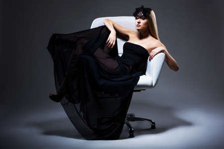 Enjoyment. Classy Elegant Woman Blonde relaxing in Chair. Black Dress and Mask with Feathersの写真素材