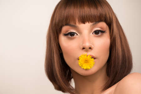 Face close up. In the mouth holds a dandelion flower. Arrows, long eyelashes, makeup. Beauty portrait. Cleansing healthy skin. Hairstyle with bangs.の写真素材