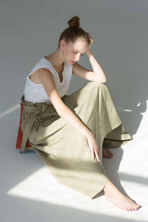 Young woman posing in studio on a light background. Long green skirt, white summer T-shirt, black sandals. She stands sideways, hands in pockets.の写真素材