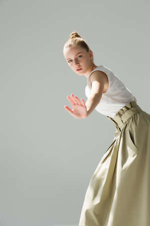 Young woman posing in studio on a light background. Long green skirt, white summer T-shirt, black sandals. She stands sideways, hands in pockets.の写真素材