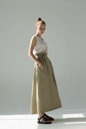 Young woman posing in studio on a light background. Long green skirt, white summer T-shirt, black sandals. She stands sideways, hands in pockets.の写真素材