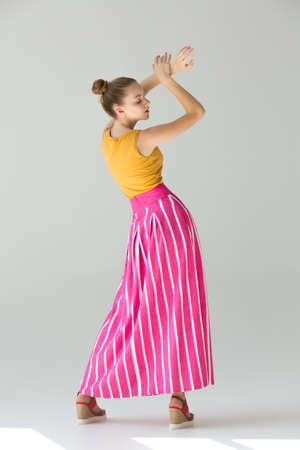 The girl stands with her back in full growth, her arms are raised above, her eyes are closed. Clothes - long red skirt in wide white striped and orange tank top. Summer look.の写真素材