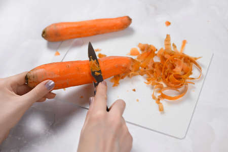 Female hands peel carrots with a knife on a white board on a light table.Cleaning fresh carrots before serving and cooking.Peels carrots from the peel. high quality photoの写真素材
