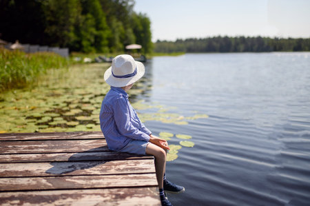 A boy in a hat sits on a wooden pier by the lake on a summer day. The boy dreams looking into the distance. Copy space.の写真素材