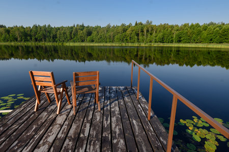 Two wooden chairs on a wooden jetty overlooking a calm lake and forest on a sunny day. The concept of relaxation,の写真素材