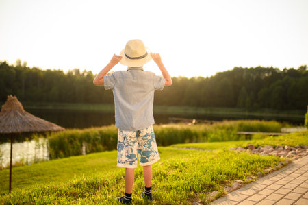 A boy in a hat looks into the distance at the lake at sunset,enjoys the view of the calm lake and the forest, breathes fresh clean air.Vacation, summer concept.の写真素材