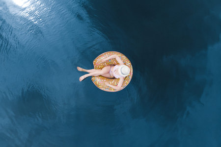 Aerial view of a young woman in a swimsuit, a hat, floating on an inflatable big donut in the...