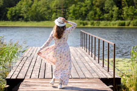 A woman in a hat and dress walks on a wooden pier near a lake and forest on a summer dayの写真素材