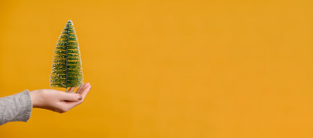 Womans hand is holding a mini Christmas tree on a yellow background.Banner.Concept of the new year,creative ideasの写真素材