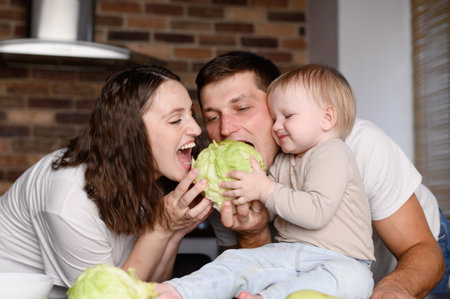 While preparing a healthy meal in kitchen, child holds a cabbage head in his hands, and mom and dad take turns biting it. Healthy eating,organic vegetables, plant-based diet. Proper nutrition conceptの写真素材
