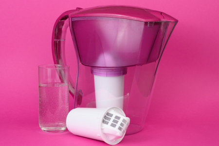 Water purification jug with spare filter and a glass of purified drinking water on pink background. Concept of water purification, drinking regimeの写真素材