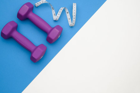Layout of two rubberized dumbbells 2 kg purple, measuring tape on a blue and white background, top...