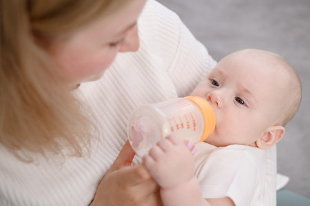 Mom holds newborn baby in her arms, baby drinks adapted milk formula from bottle held by mother,...