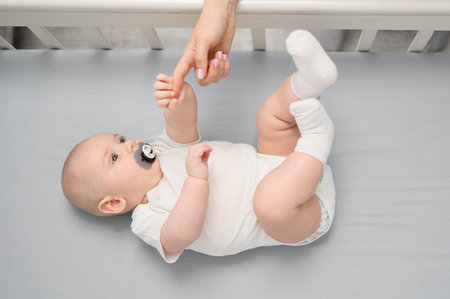 Top view of a newborn with a pacifier lying in crib, mom gave her hand to her infant so that he felt...