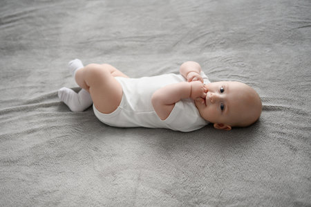 Adorable newborn baby in white bodysuit lies on his back in crib in children room and sucks his fingers. Child explores parts of his body using taste sensations. Baby care concept, baby productsの写真素材