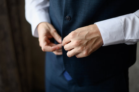 Fashionable man buttoning button on his vest while standing indoors, close-up. Wedding day, grooms morning. Young modern businessman, mens fashion and mens clothing styleの写真素材