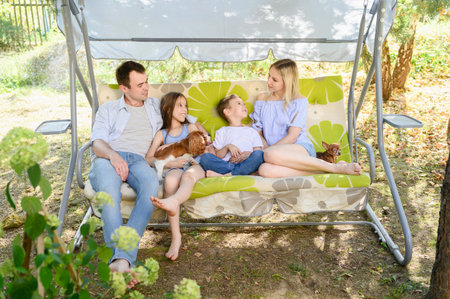 Family swings on large garden swing in backyard of country house. Parents with boy and girl sit on swing, talk, pet their favorite pets. Concept of country summer vacation, weekendの写真素材