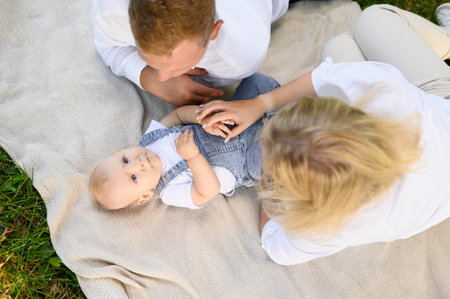 Parents relaxing in park on weekend, lying on blanket next to baby. Toddler is lying on his back, mom and dad are tickling child, enjoying time together, stroking him, top view. Happy family conceptの写真素材