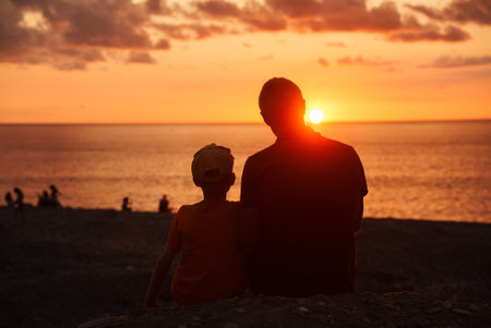 Back view of dad , son sitting at sunset on seashore enjoying summer sunset. Tourists sitting on beach looking at setting sun. Travel concept, seaside vacation, rest, leisure time of child and fatherの写真素材
