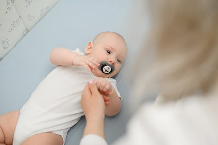 Cute little newborn baby lies in cozy bed, happily sucks pacifier, looks at his mother leaning over...