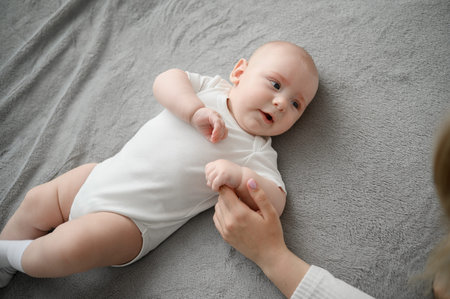 Cute little baby lies on soft bed, caring woman holds tenderly by the hand, creating warm...