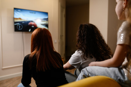 Back view of women, child sitting on sofa in living room playing game console racing with joystick controller spending leisure time together. Cute family having fun on weekend playing video gamesの写真素材