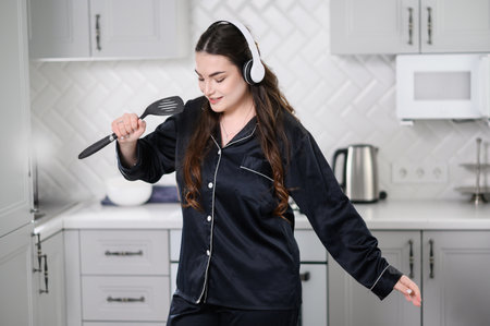 Woman singing and dancing in kitchen at home. Happy girl holding spatula as microphone, listening to music on headphones while cooking, having fun with kitchen utensils. Cooking concept at homeの写真素材