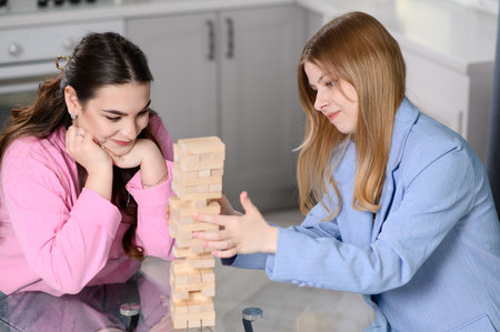 Two friends are sitting in living room, chatting, spending their free time playing board game Falling Tower game on weekend. Girls take turns removing wooden blocks from tower, keeping it from fallingの写真素材